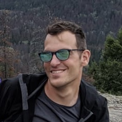 Shay Elkin, a man with short brown hair, is smiling at the camera. He is outdoors, on a rocky overlook with a forested, mountainous landscape blurred in the background. He is wearing a dark jacket and reflective sunglasses.